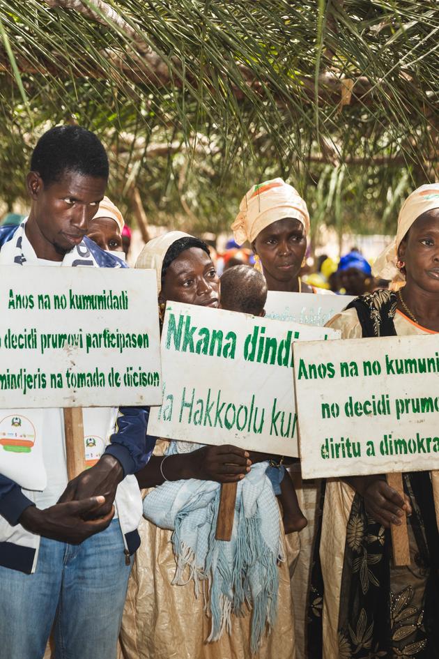 في ظل أومار كبير في أوراق النخيل ، يلوح القرويون بعلامات في لغات ماندينغو و Peule و Creole لتأكيد رغبتهم في إنهاء تشويه الأعضاء التناسلية للإناث داخل مجتمعهم.