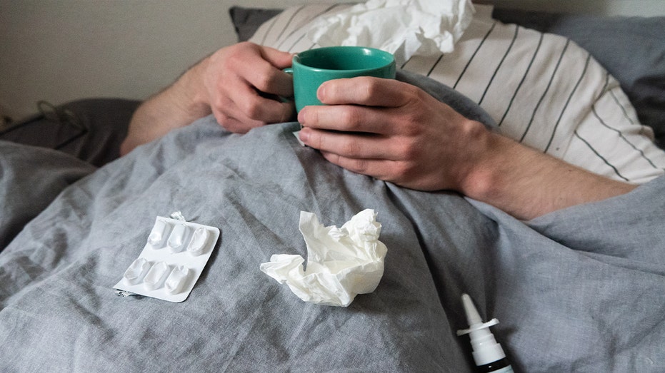 رجل يرقد في السرير ومعه مناديل وفنجان شاي ورذاذ أنف وأقراص.