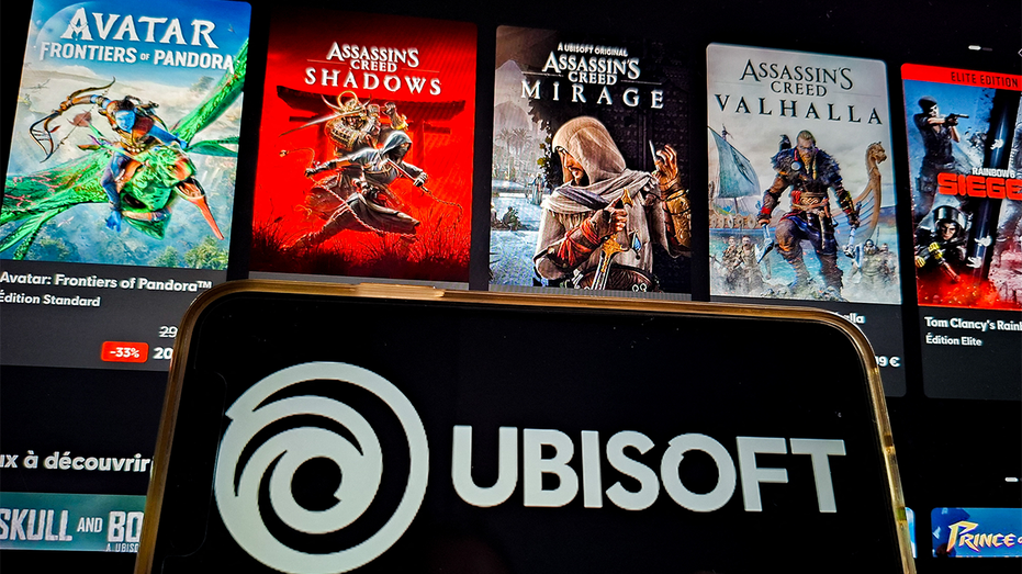 رسم توضيحي من Ubisoft في فرنسا، 27 يناير، 2026. هاتف ذكي يعرض شعار Ubisoft بشكل حلزوني أرجواني وحروف سوداء، أمام ثلاثة ملصقات ترويجية لسلسلة Assassins Creed.
